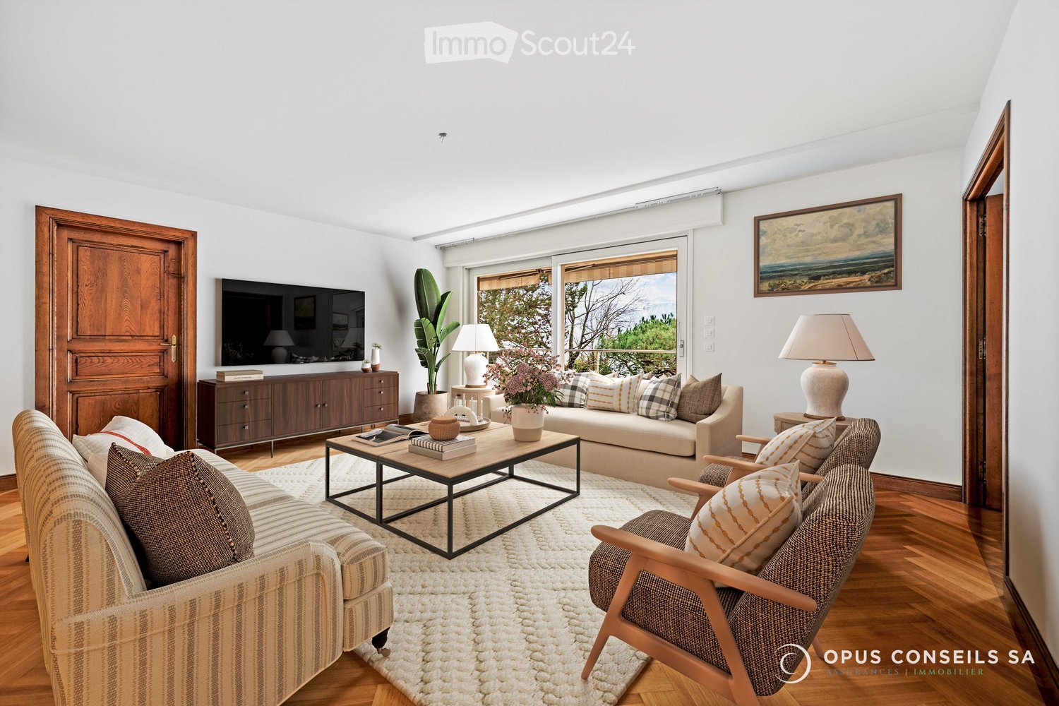Bright living room with wooden flooring, large windows, and a view. Features a beige sofa, coffee table, armchair, table lamps, and a TV on a wooden cabinet.