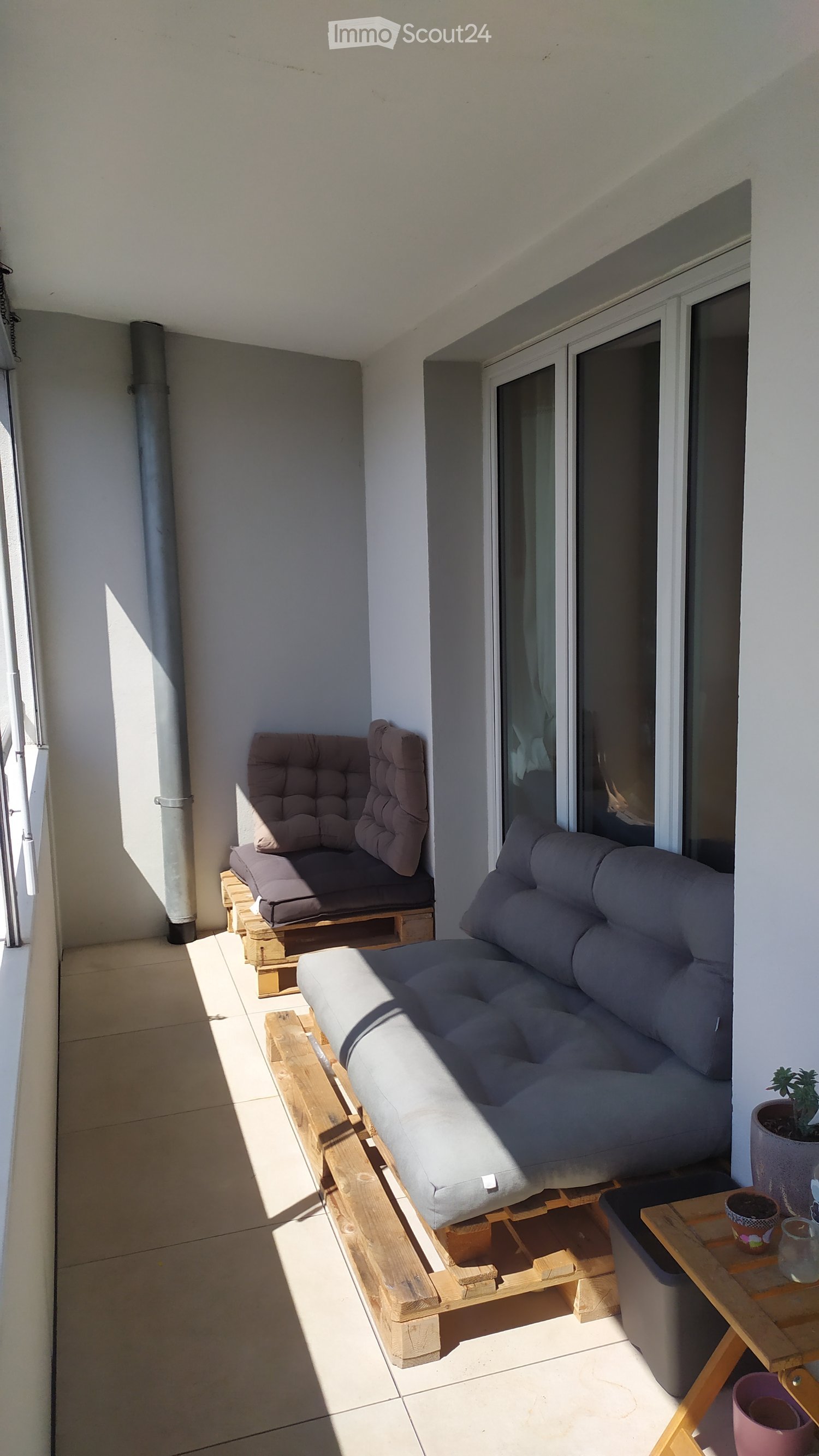 Balcony with a grey sofa, wooden chair, small table, and potted plants.
