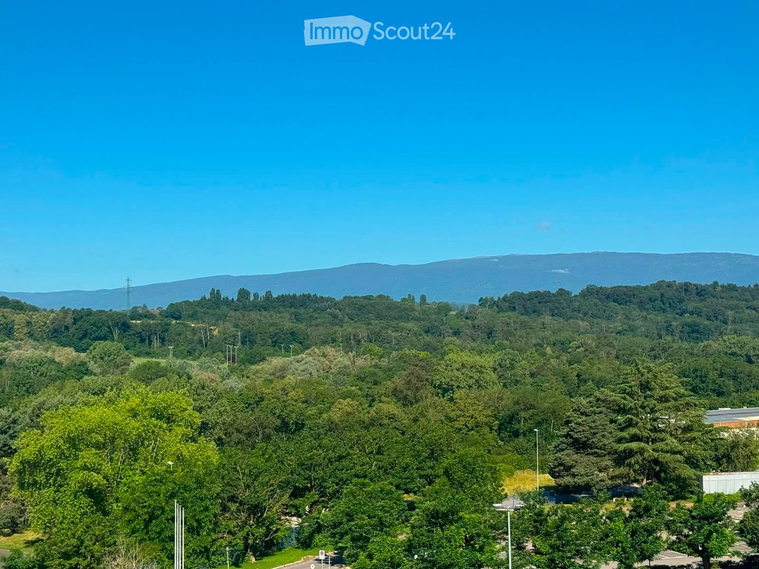 A panoramic view of a lush, green landscape with dense forest covering the lower hills and a clear blue sky above. The area includes a mix of trees, open spaces, and a few scattered buildings in the distance.