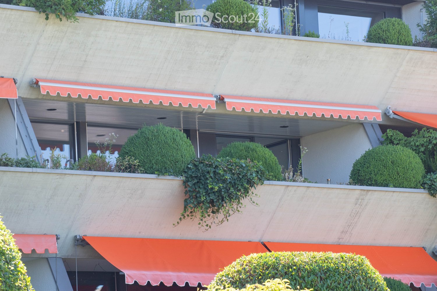 Multi-story building with balconies, red awnings, and greenery including shrubs and climbing plants.