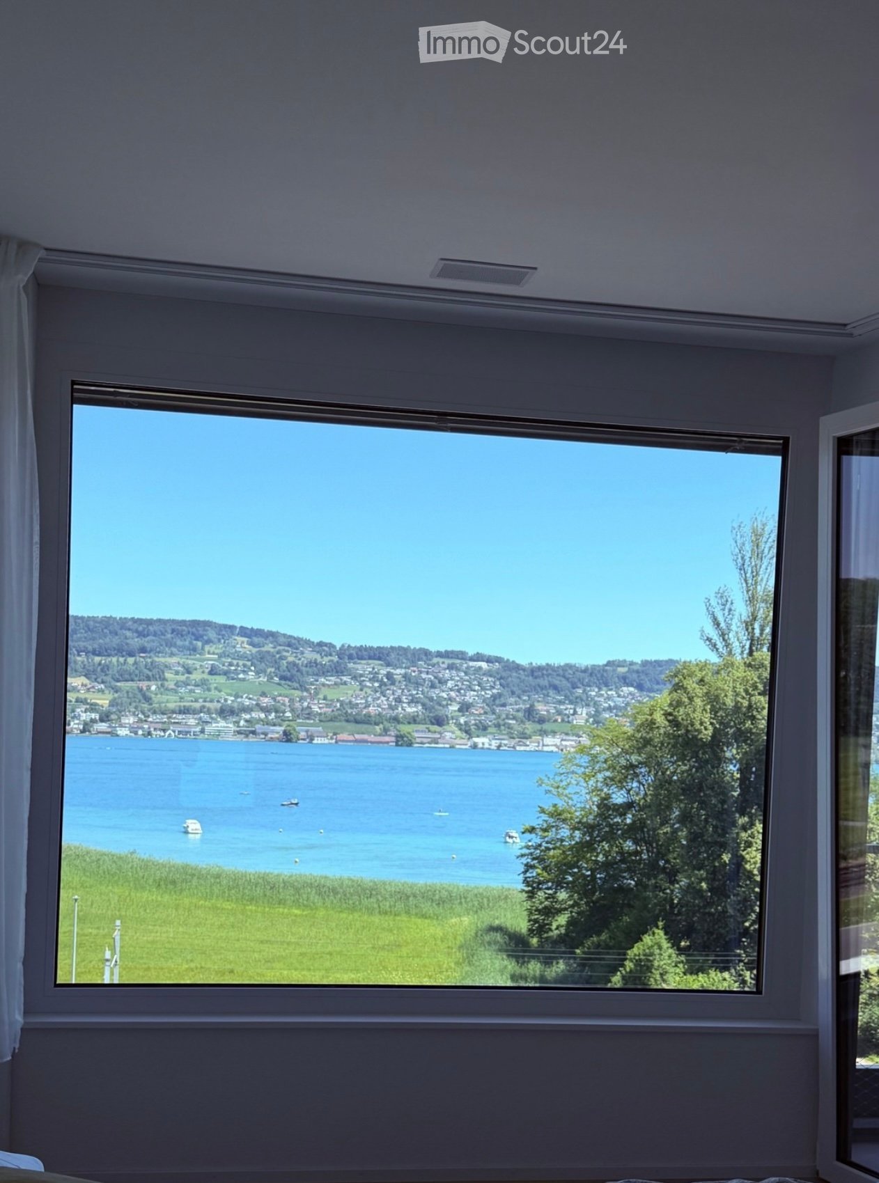 Empty room with large window offering a scenic view of a lake, surrounding greenery, and distant town.
