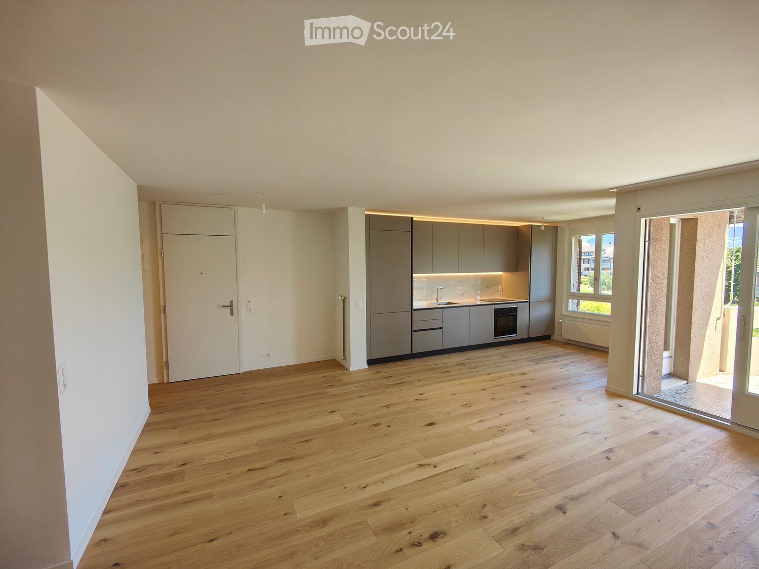 Empty room with light wooden flooring, white walls, grey kitchen cabinets, and a large window with a view of greenery.