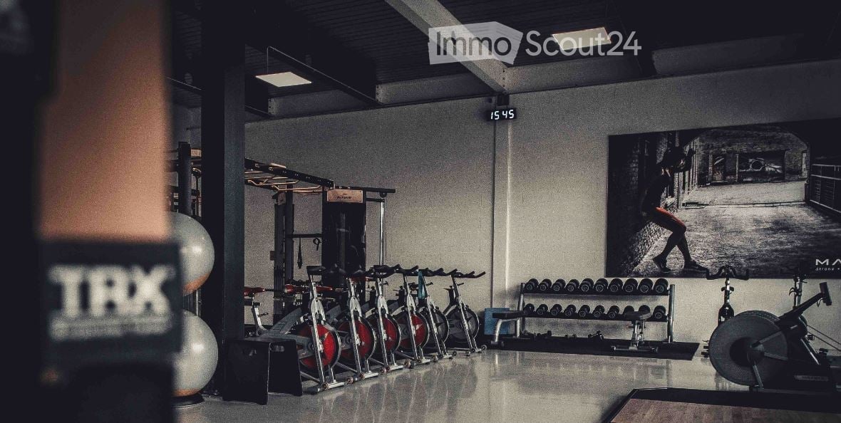 Modern gym space with multiple exercise bikes, dumbbell rack, fitness ball, and weight machine. Large black-and-white poster on wall.