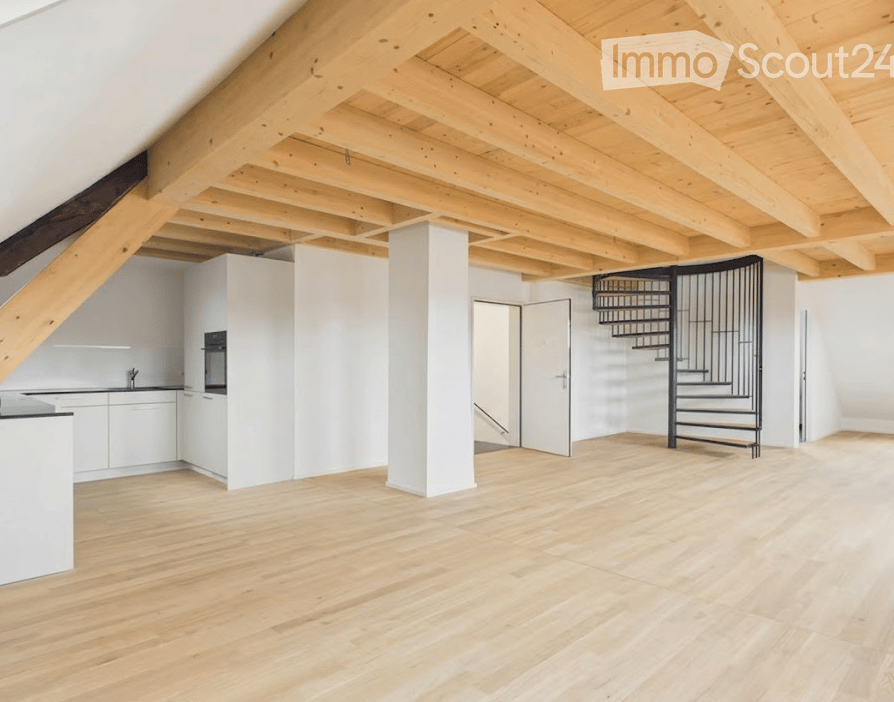 Bright, open loft space with wooden floors and exposed wooden beams. Features a modern white kitchen area with cabinets and a sink, a black spiral staircase leading to an upper level, and large white walls.
