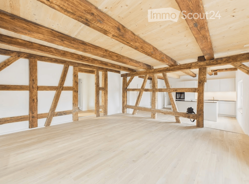 Empty room with exposed wooden beams and light wooden flooring, white walls, and a small kitchen area visible in the background.