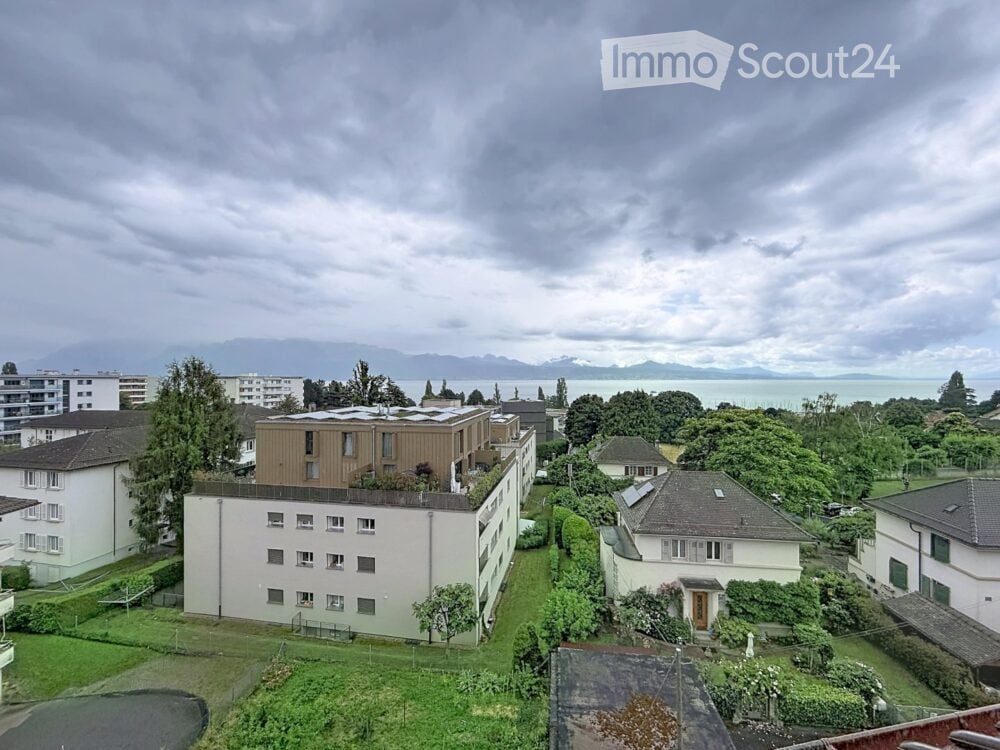 A multi-story residential building with a flat roof and modern design, surrounded by greenery and other buildings, with a scenic view of a lake and mountains in the background.