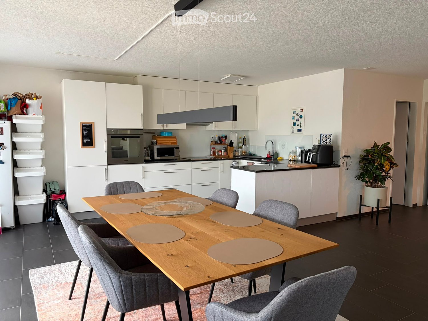 Modern kitchen with white cabinets, a wooden dining table with six chairs, a refrigerator, oven, microwave, and a potted plant in the corner.