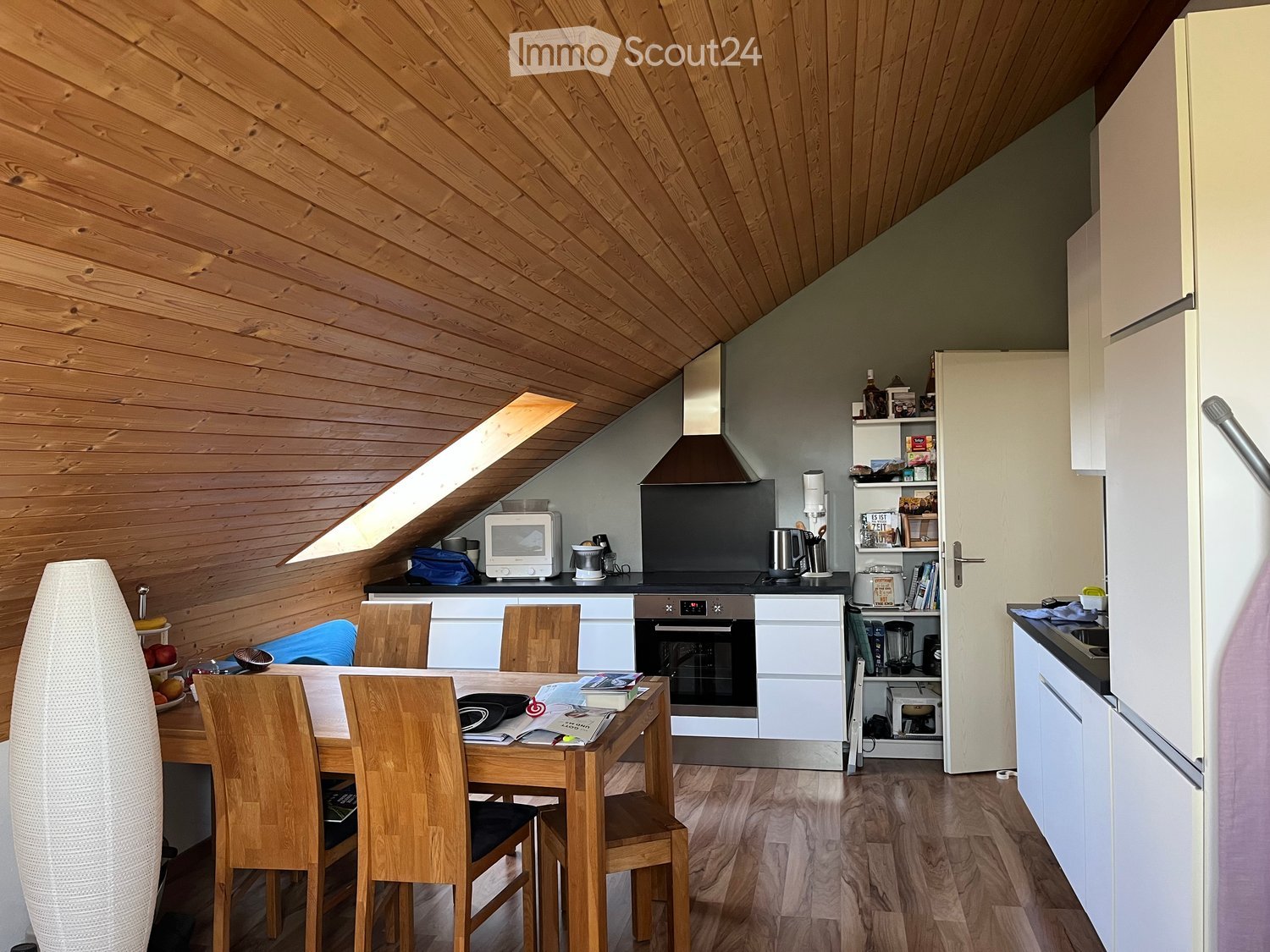 Modern kitchen with wooden ceiling and walls, white cabinets, stainless steel appliances, dining area with wooden table and chairs, and built-in shelving unit.