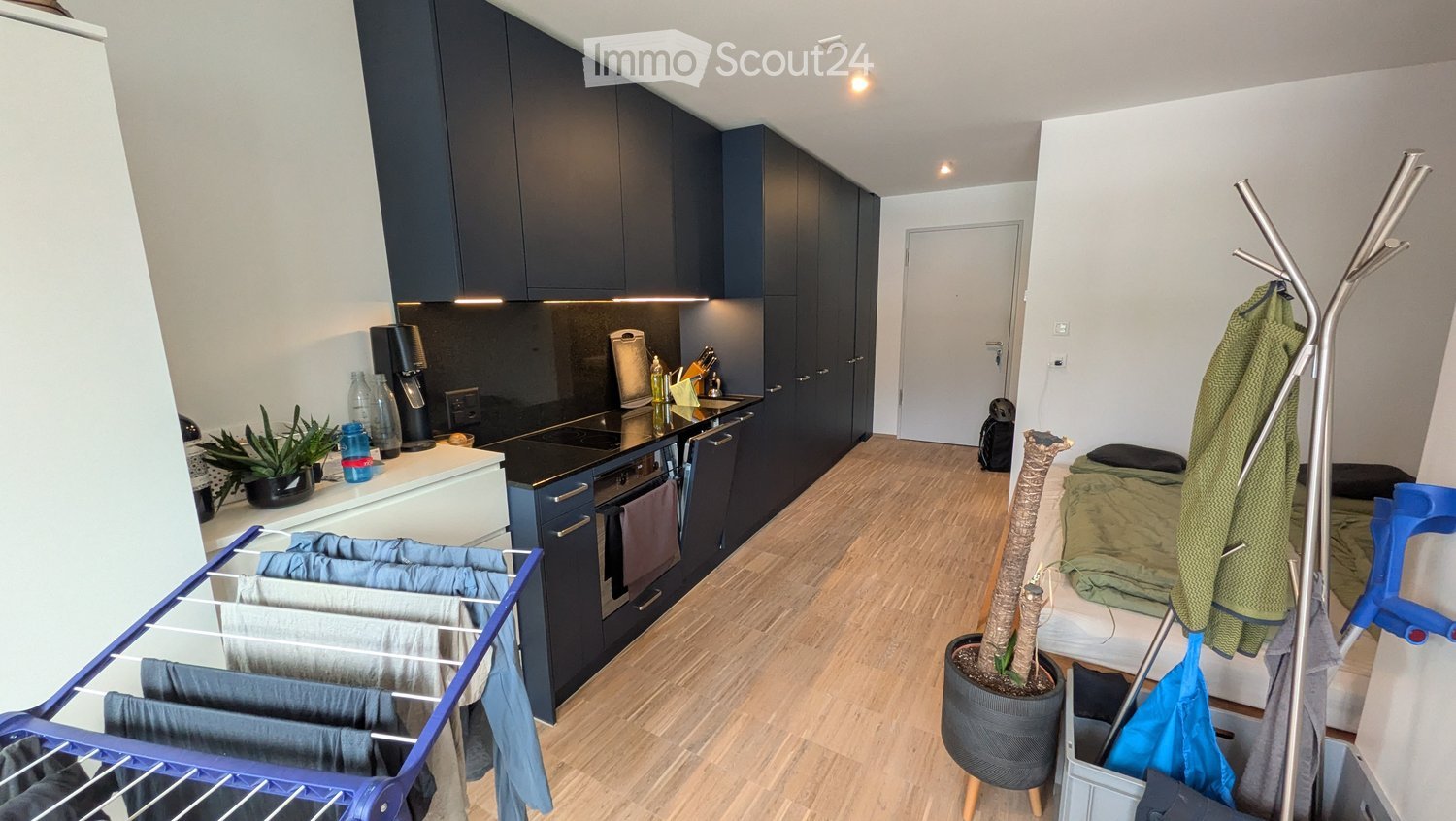 A compact studio apartment with a bed, a small kitchen area featuring a fridge, oven, and sink, and a clothes drying rack. A coat rack and a potted plant are also visible.