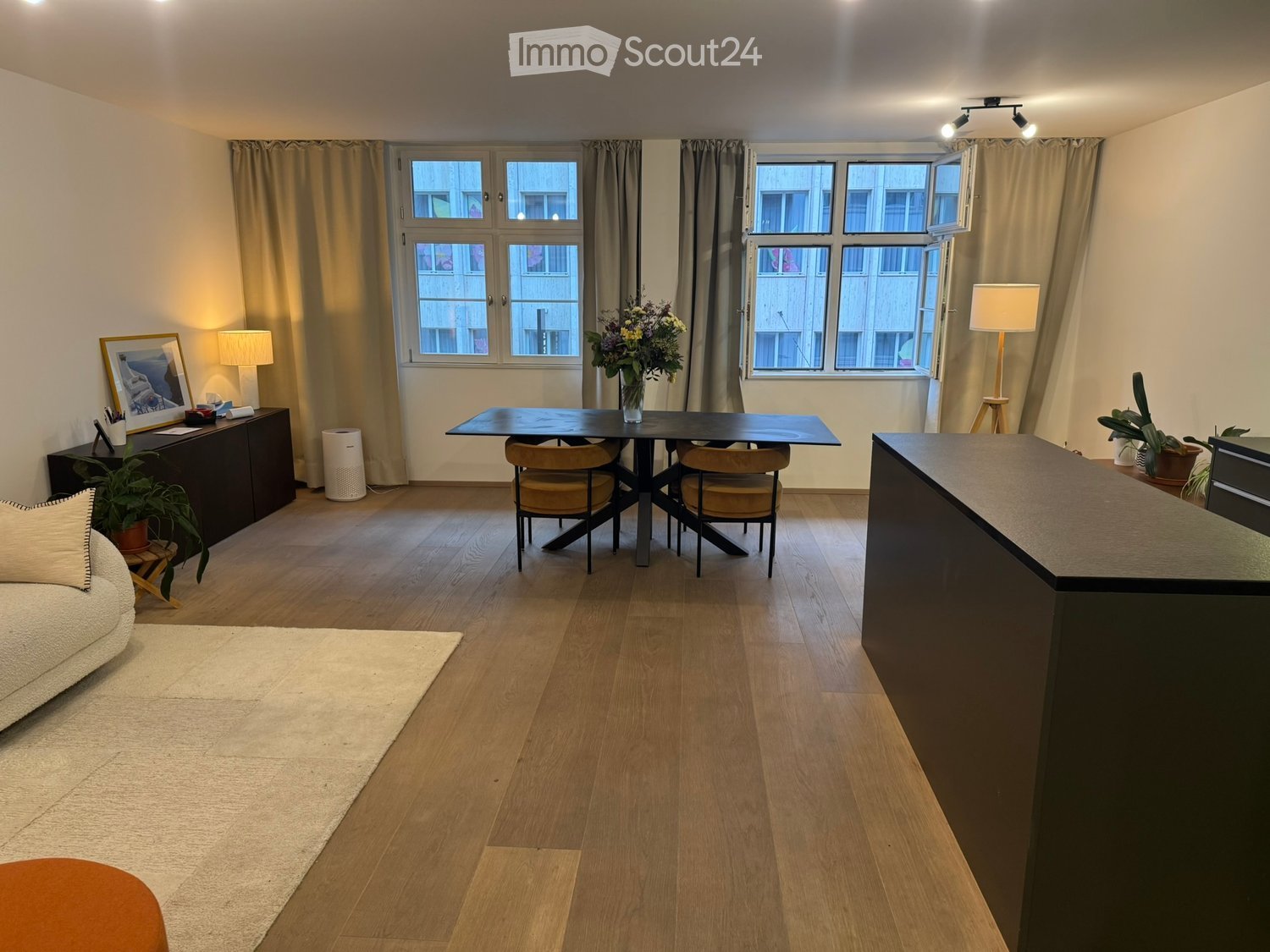 Open-plan living and dining area with wooden flooring, large windows with curtains, a sofa, a stool, a dining table with chairs, a cabinet, a flower vase, lamps, a picture frame, and plants.