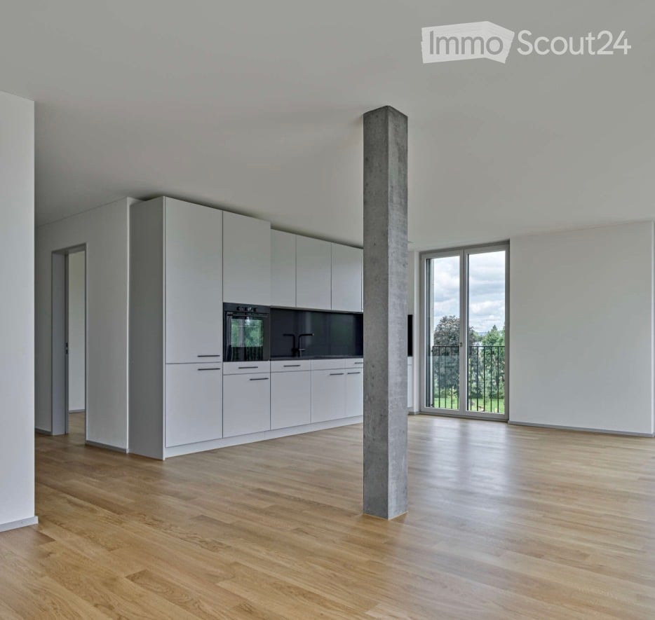 Empty kitchen with white cabinets, wooden flooring, and a large window overlooking a green area.