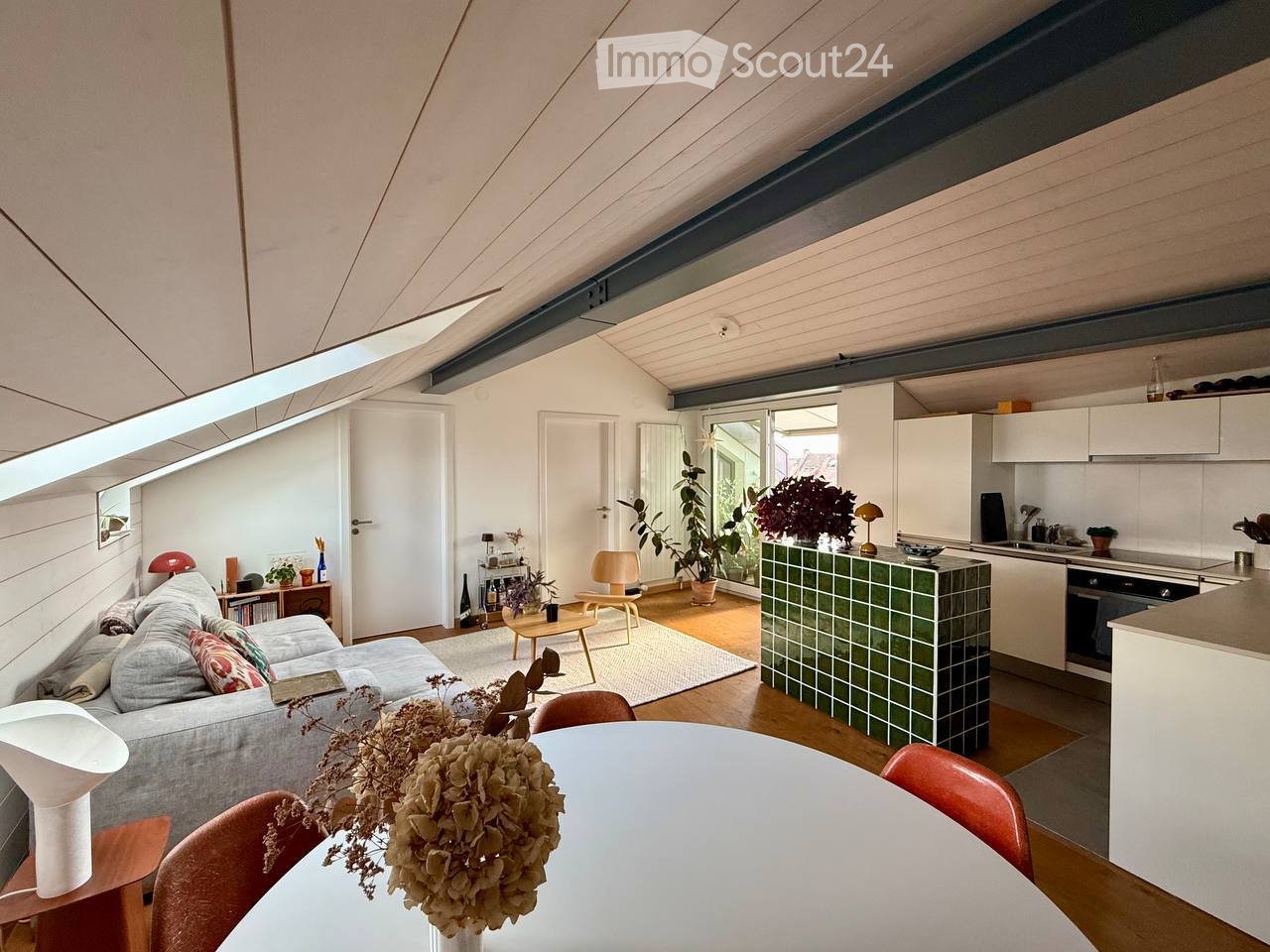 Bright, open living space with a grey sofa, coffee table, dining table with chairs, kitchen area with white cabinets and a green-tiled island, and several plants and decorative items.