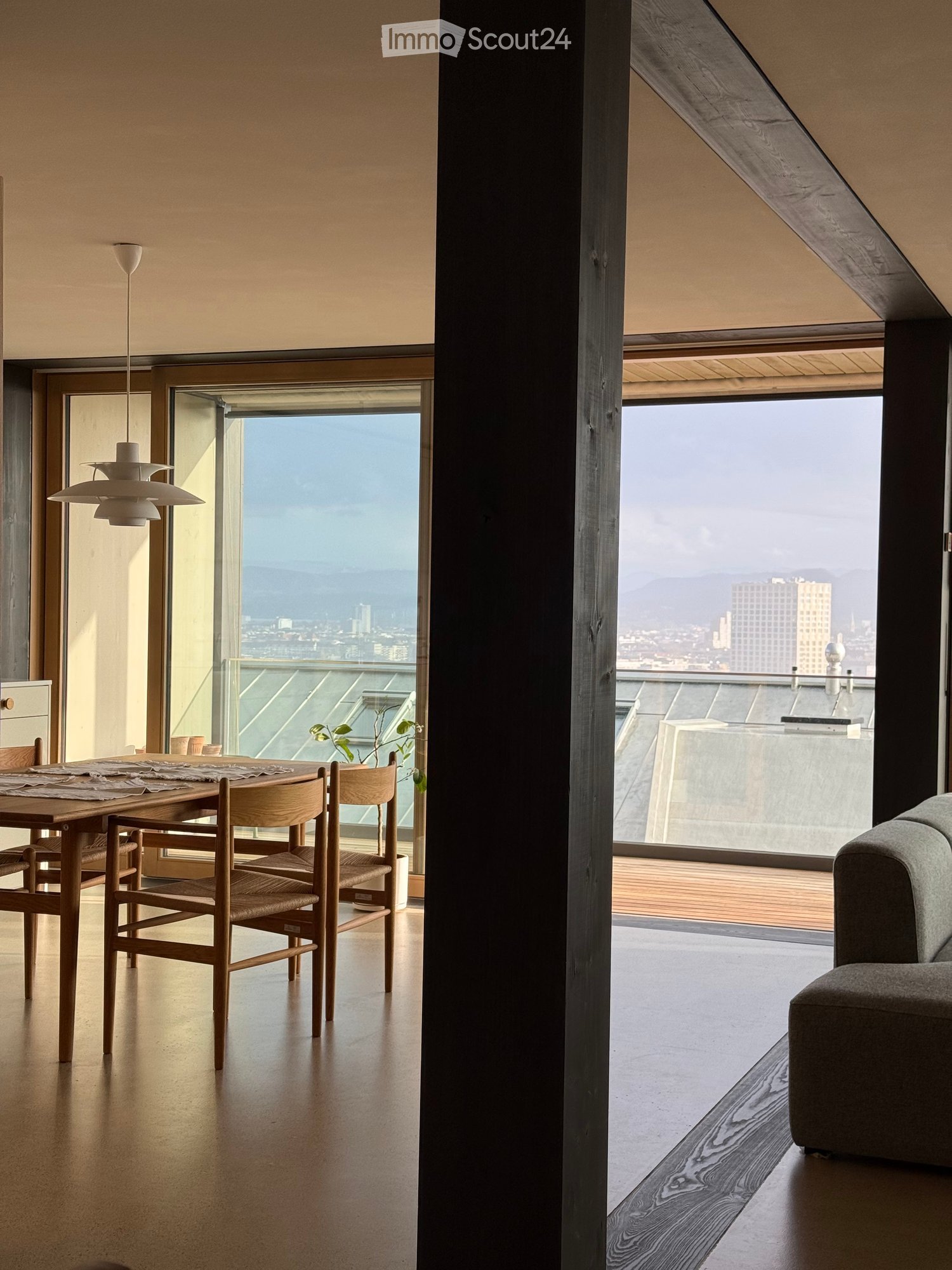 Modern dining area with a wooden table, chairs, pendant light, and a grey sofa, overlooking a cityscape with a view of mountains and buildings.
