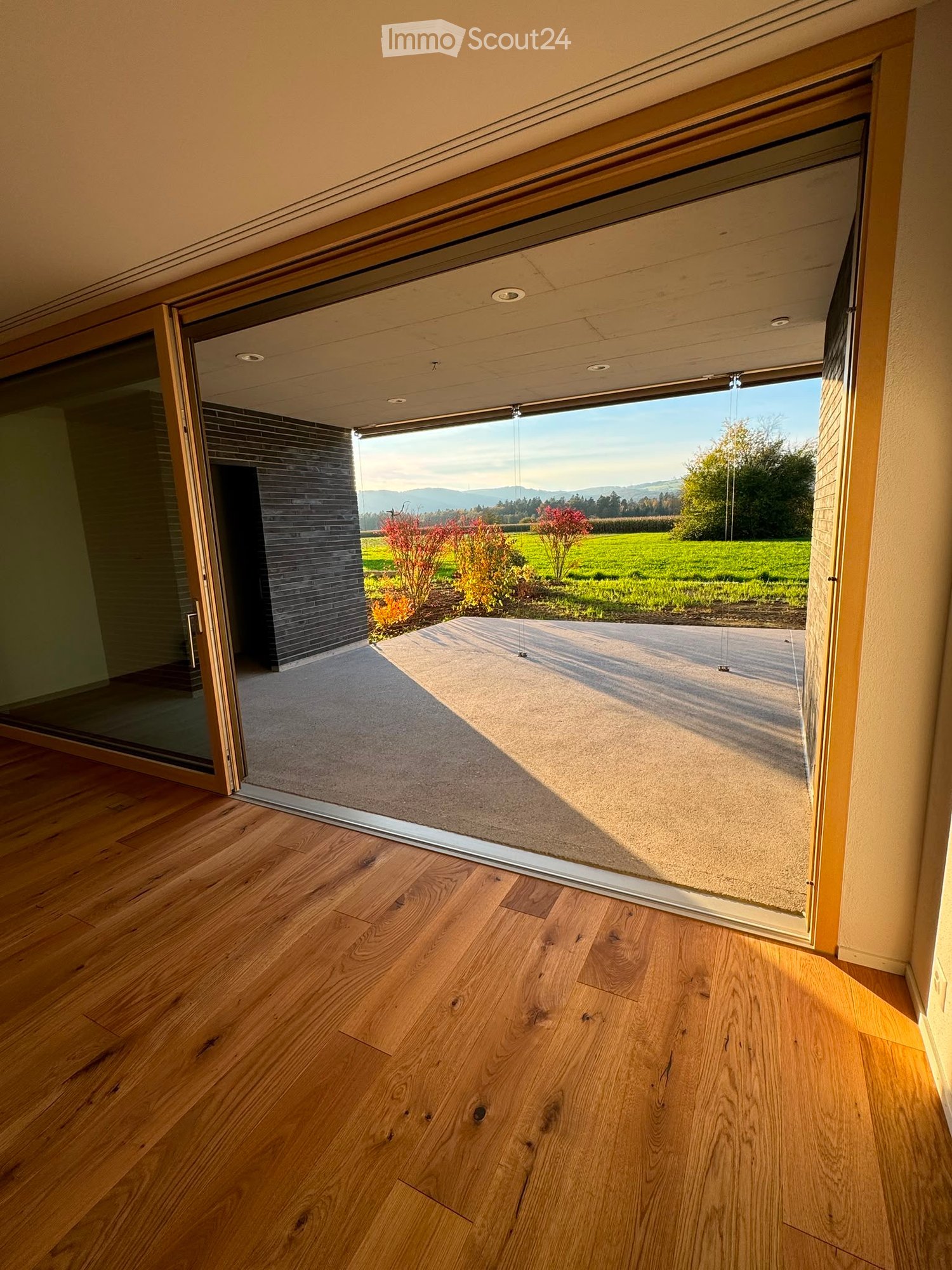Open sliding glass door leading to a patio with wooden flooring inside and a view of a green landscape outside.