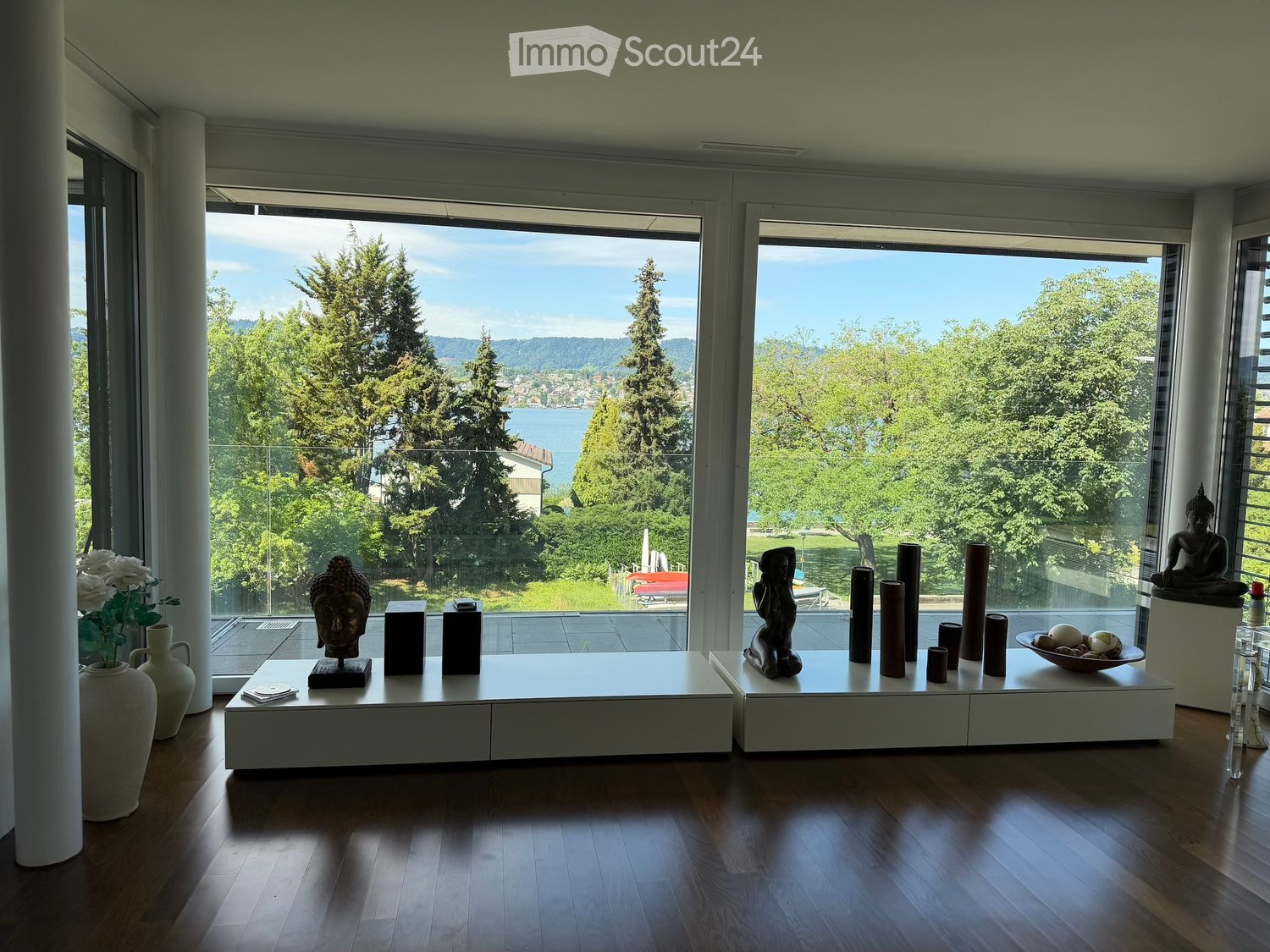 Bright living room with large windows offering a scenic view of trees and water. Decor includes white tables, vases, sculptures, and a bowl with decorative items.