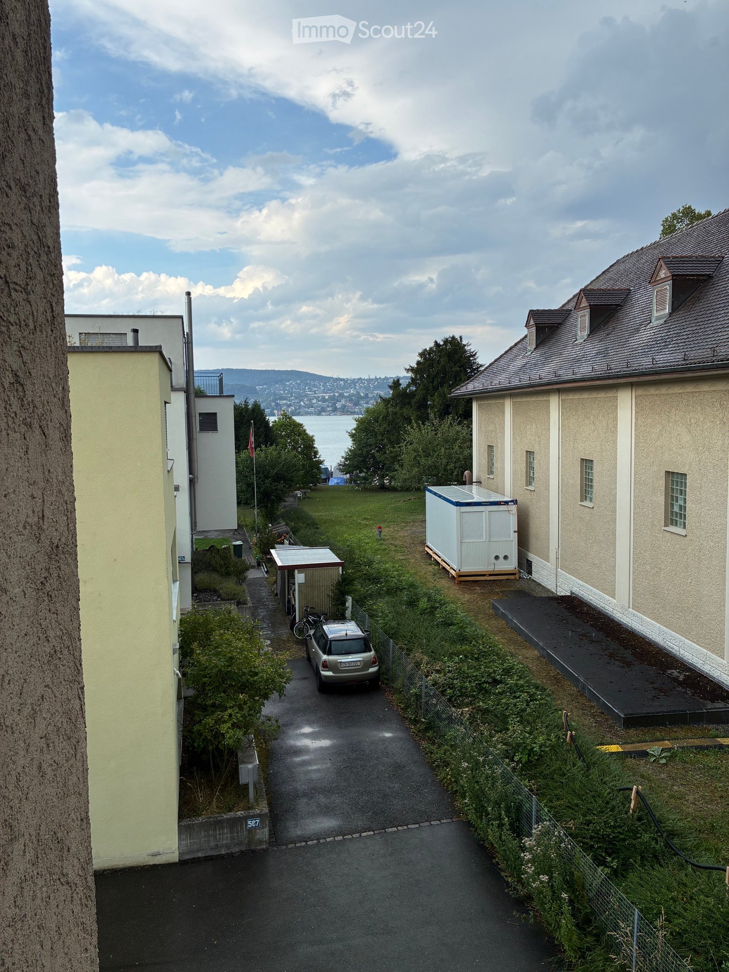 A narrow alleyway between buildings with a parked car, a bicycle, and a small shed. The area is surrounded by greenery and has a view of a body of water in the distance.
