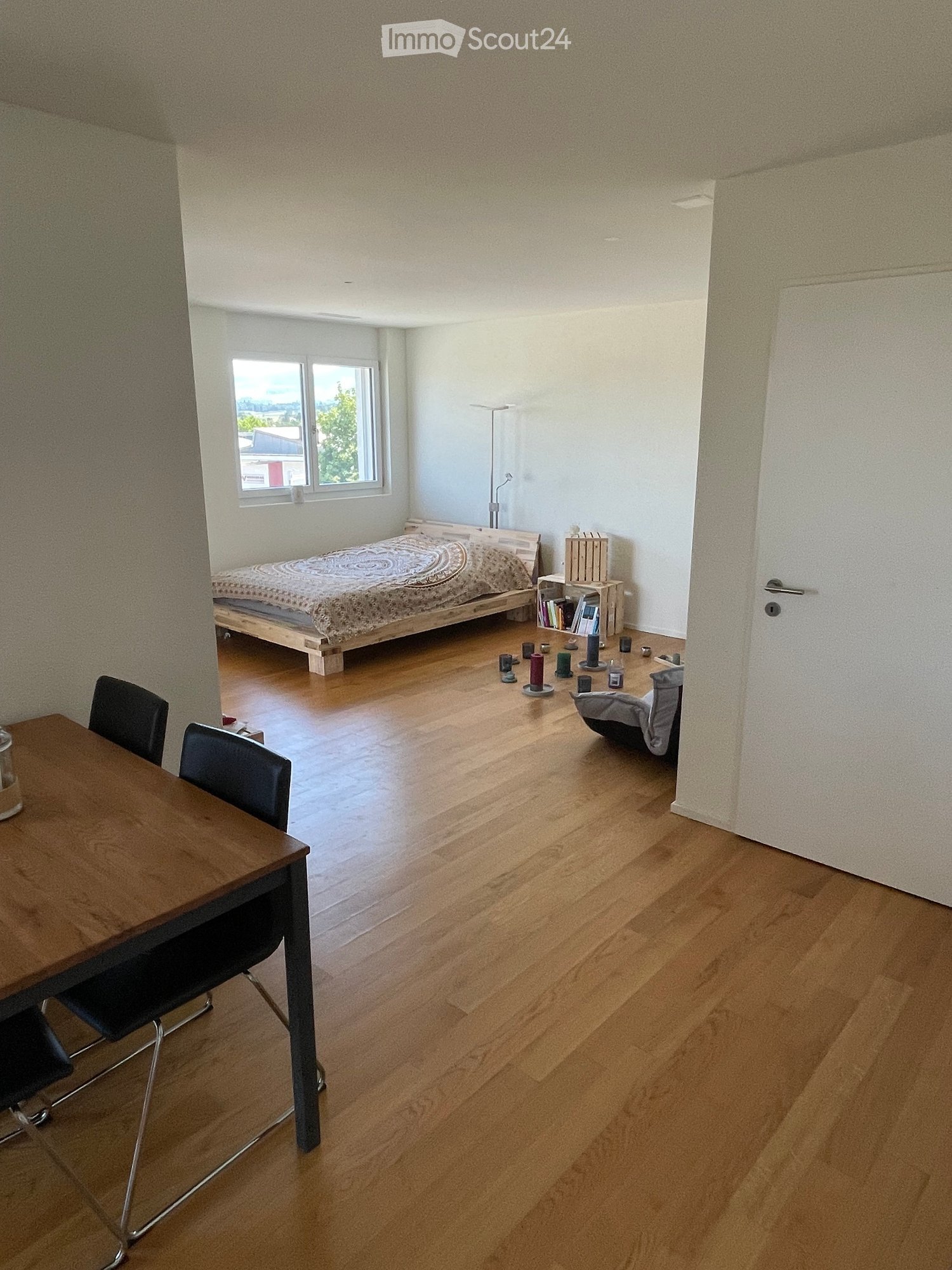 A well-lit bedroom with wooden flooring, a bed with patterned bedding, a small table with chairs, a bookshelf, and a few decorative items.