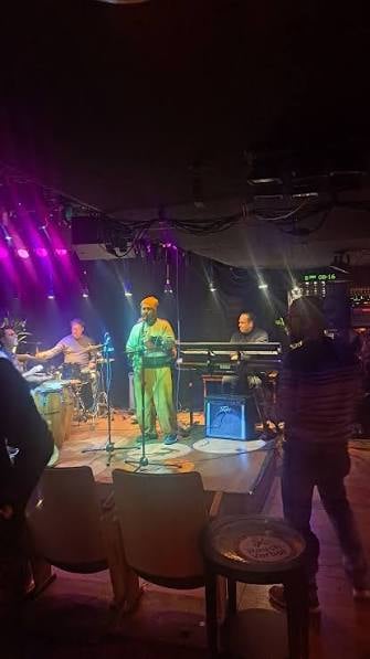 A live music performance on a small stage with a band setup, including drums, a guitar, microphones, and speakers. The stage is lit with purple and white spotlights, and there are chairs and a stool in the foreground.