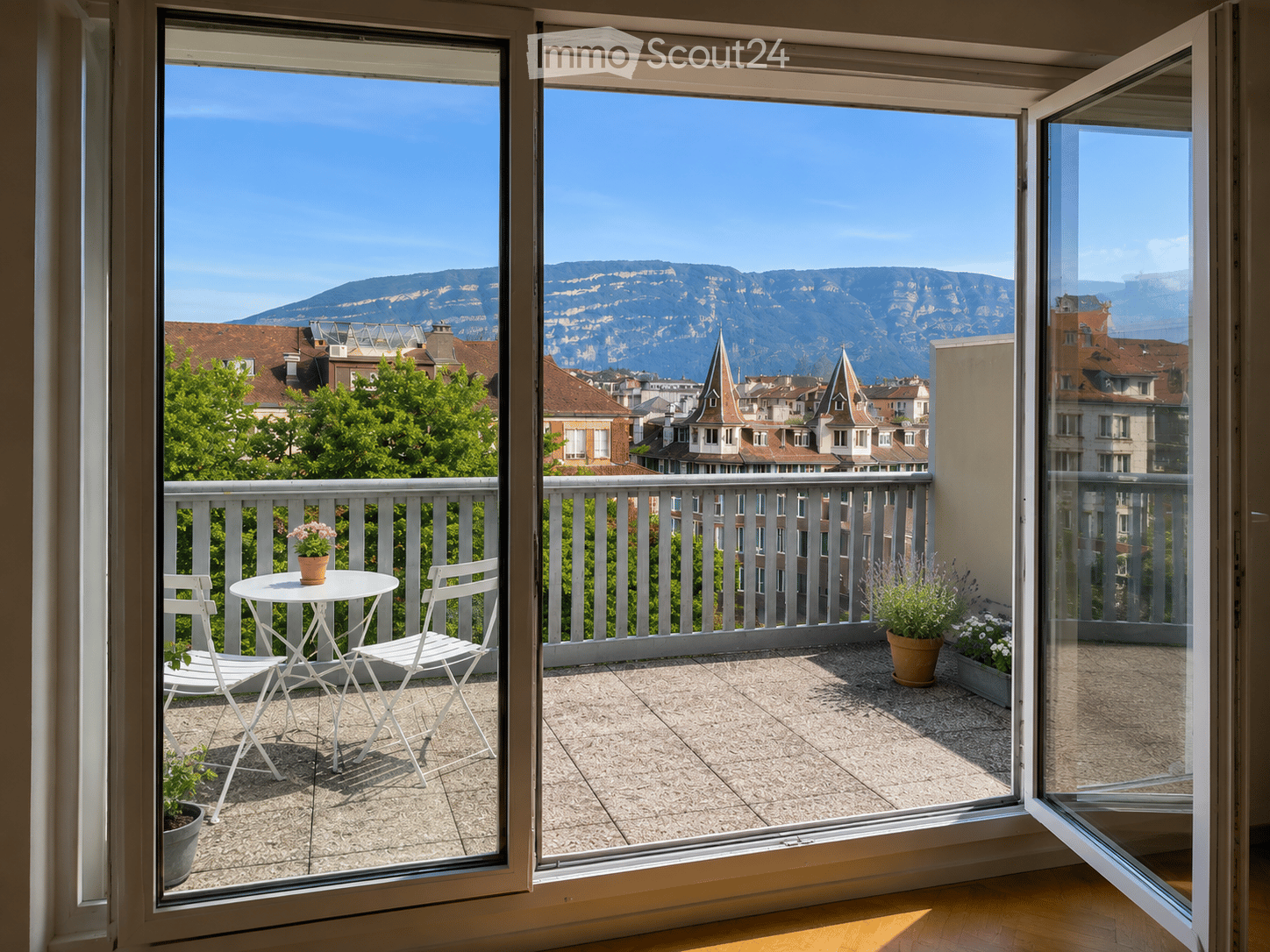 Balcony with a view of city rooftops and mountains, featuring a small table, two chairs, and potted plants.