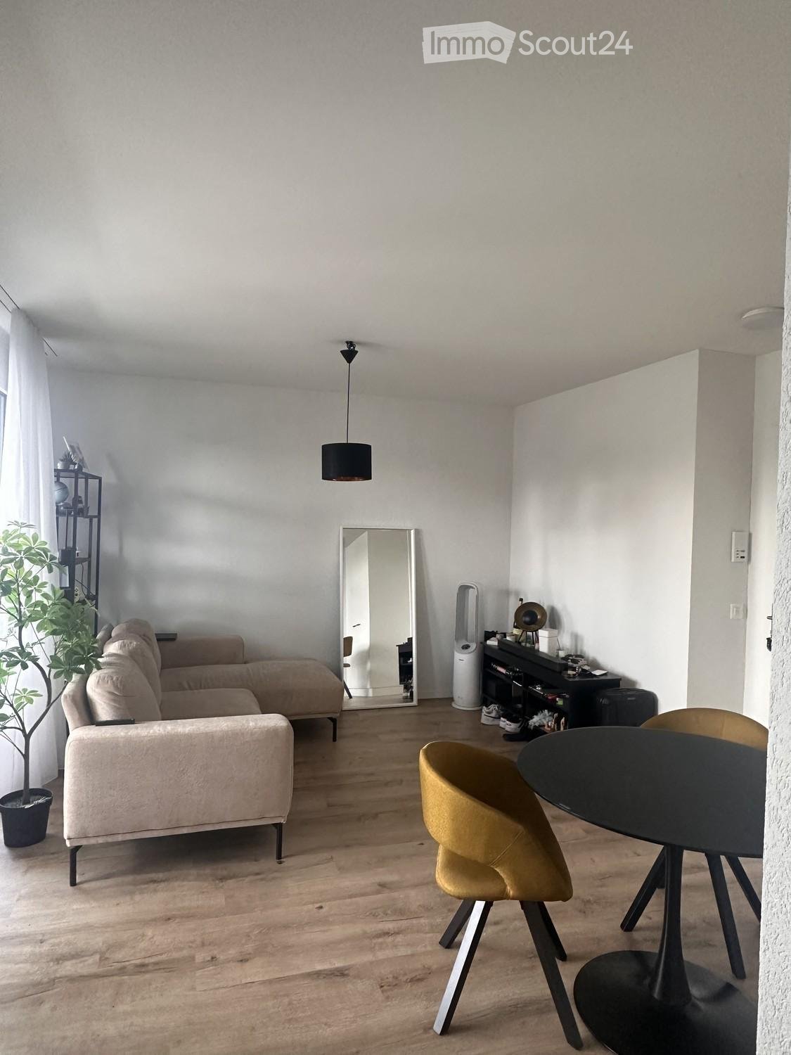 Modern living room with light wooden flooring, a beige sectional sofa, a round black dining table with yellow chairs, a large mirror, a potted plant, and a shelf with decorative items.