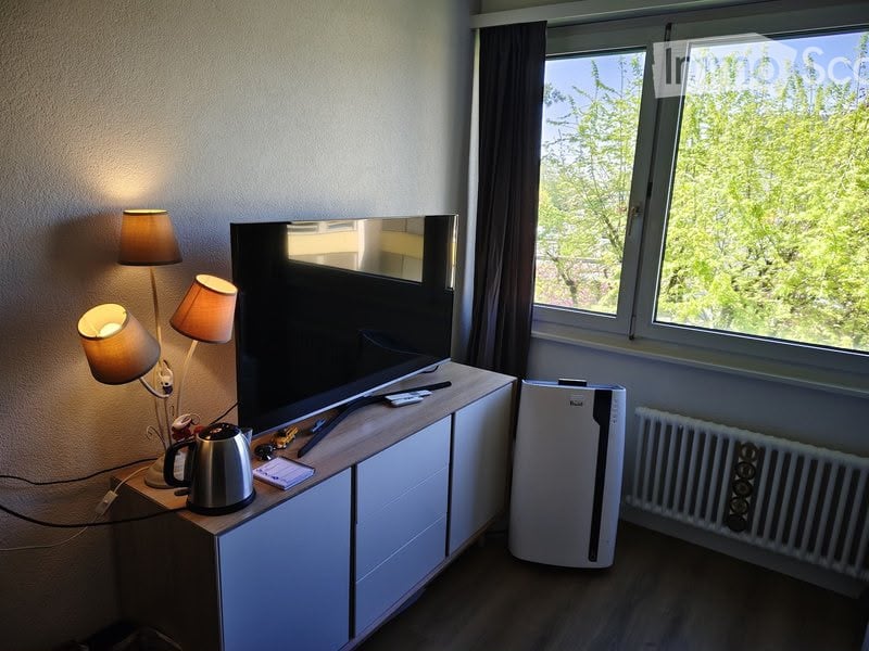 Modern furnished room with a large flat-screen TV on a white cabinet, a kettle on the table, a floor lamp with two shades, and an air conditioner unit near the window offering a view of greenery outside.