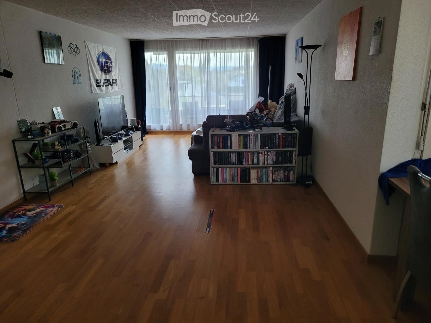 Living room with wooden flooring, large window with curtains, sofa, bookcase, shelf, and a small table with a chair.