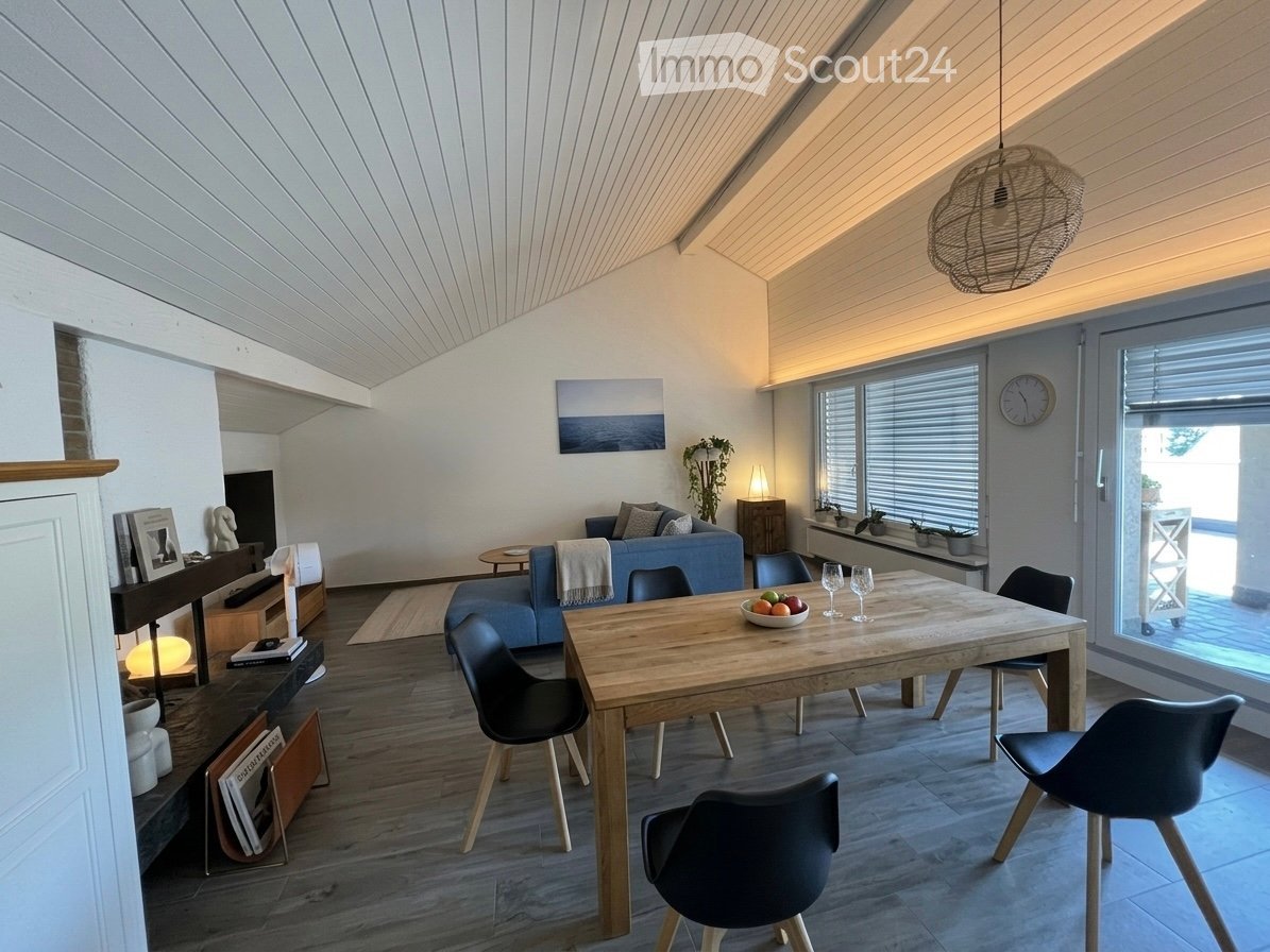Modern living and dining area with wooden flooring, white walls, and a slanted ceiling. Features a blue sofa, wooden dining table with chairs, a small side table, and decorative items including a picture frame and a clock.