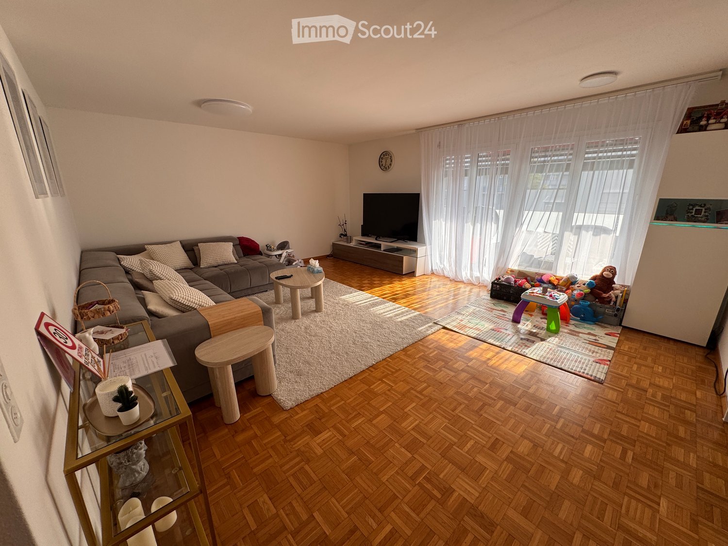Living room with wooden flooring, large windows with white curtains, grey sectional sofa, coffee table, TV stand, and various toys and decorative items.