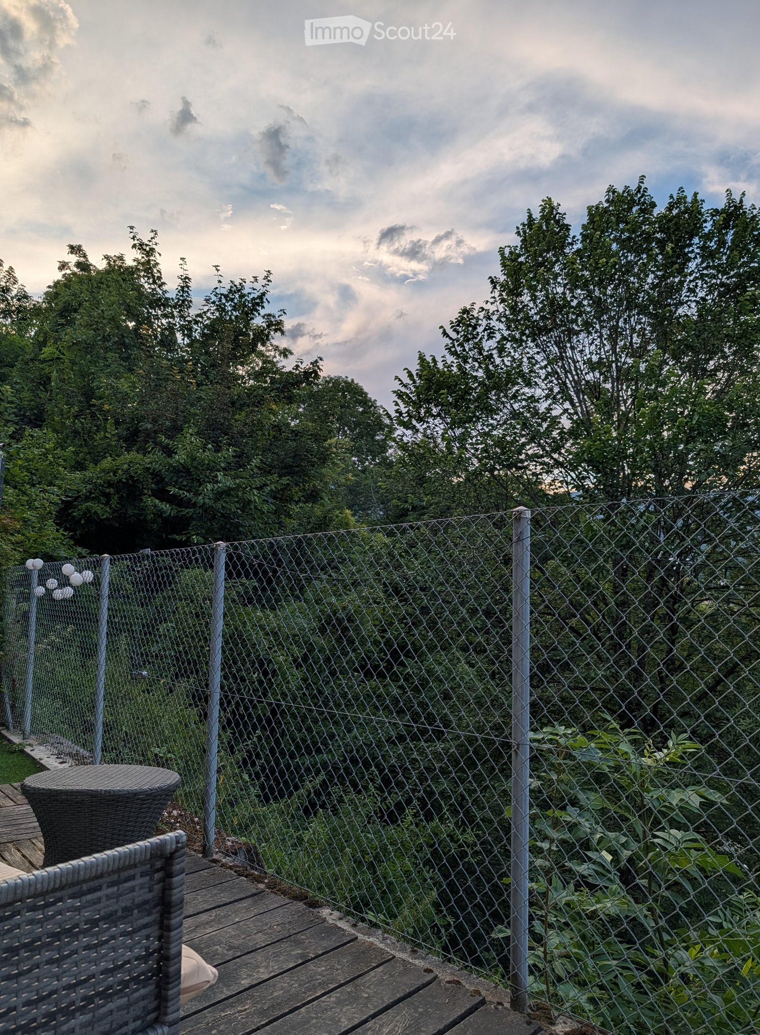 A wooden balcony with a wicker chair and table, enclosed by a chain-link fence, overlooking a lush green area with trees.