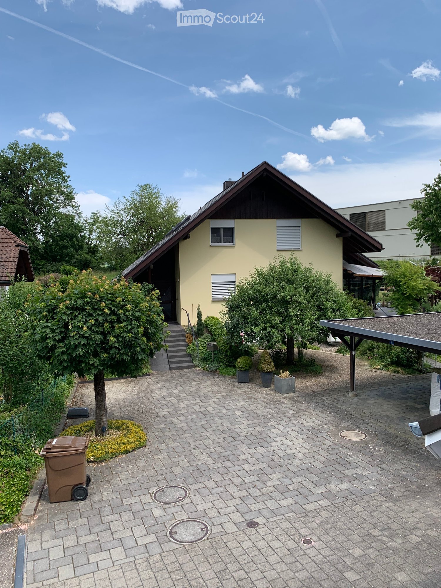 Single-story house with yellow walls, dark roof, and small porch. The entrance has steps leading up to a door, surrounded by greenery and potted plants. The driveway is paved with bricks and includes a trash bin and a covered parking area.