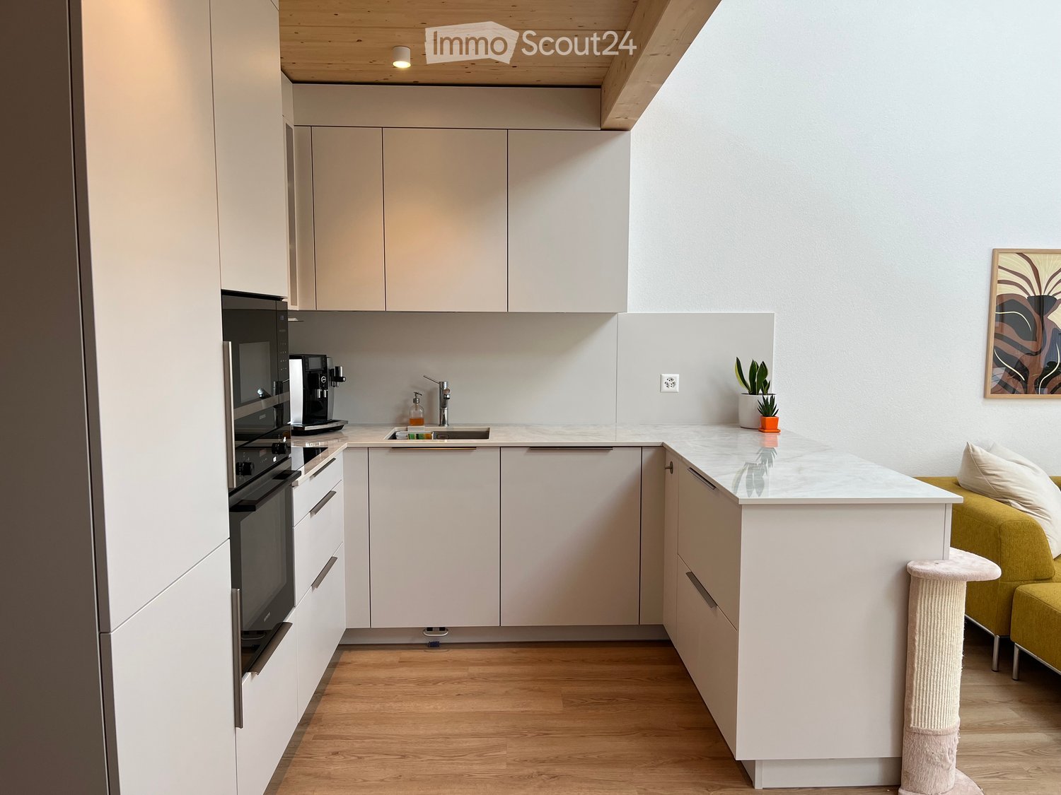 Modern kitchen with white cabinets, a central island, and a small living area with a green sofa and a cat tree. Includes a microwave, coffee maker, sink, and decorative plants.
