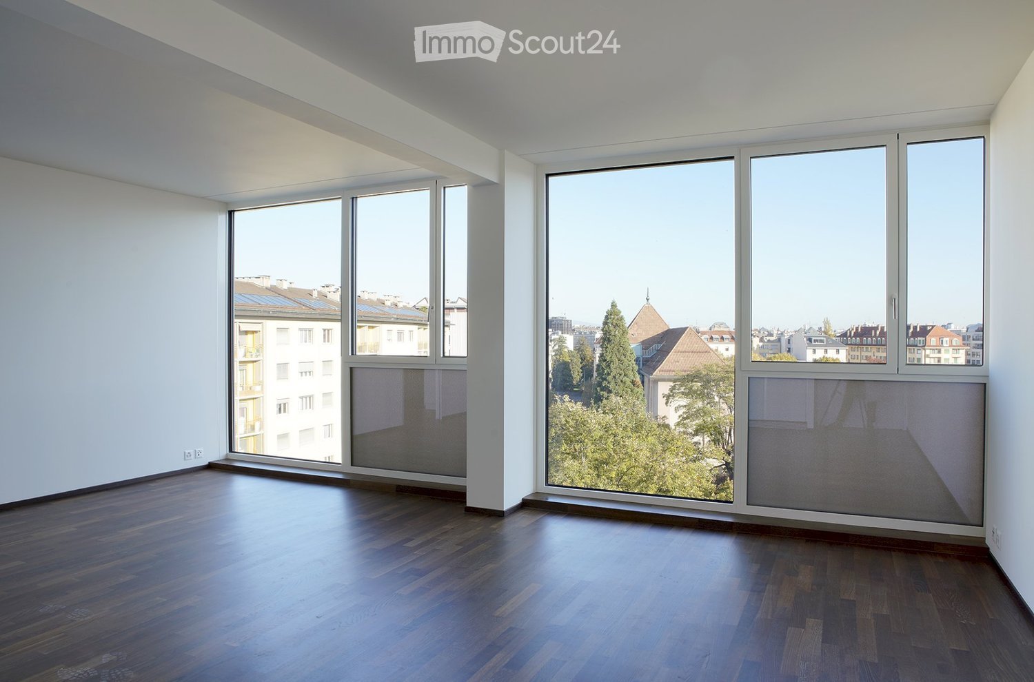 Empty room with wooden flooring and large windows offering a cityscape view. The walls are white, and the space is well-lit with natural light.