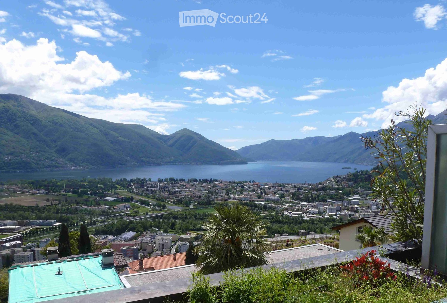 Panoramic view from a balcony or terrace overlooking a town, lake, and surrounding mountains under a partly cloudy sky.