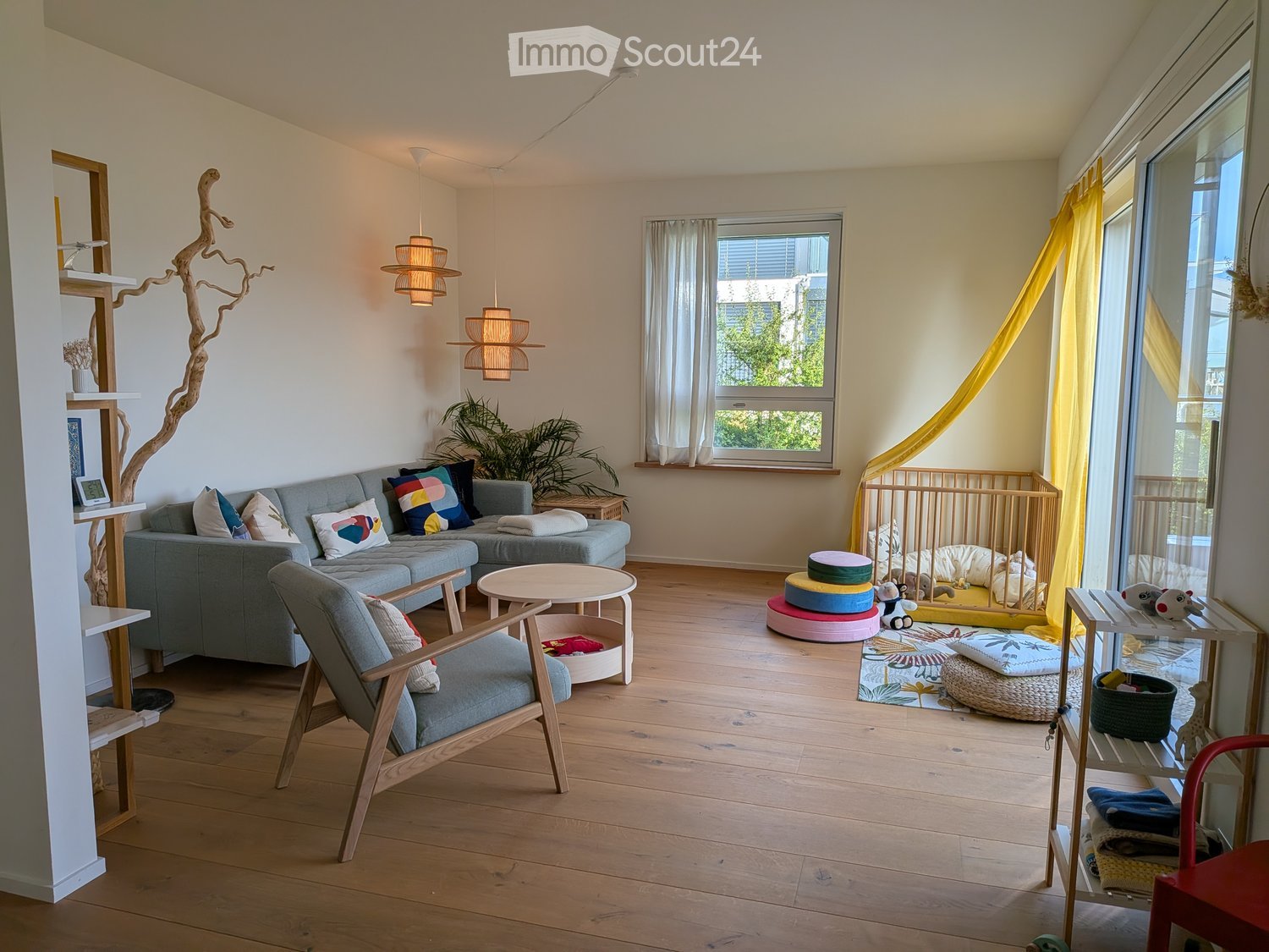 A modern living room with a grey sectional sofa, colorful cushions, a wooden chair, a round coffee table, pendant lights, a crib, and various toys.