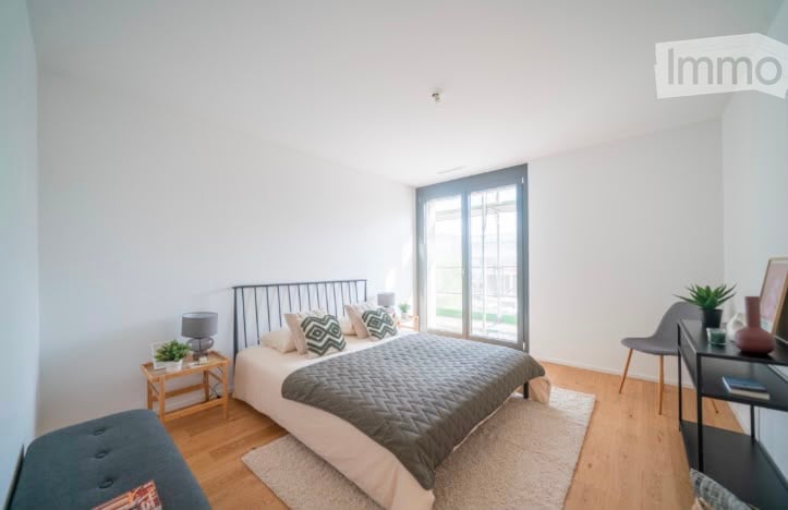 Bright, modern bedroom with a large bed, nightstand, lamp, chair, small table with plant, and picture frame on the wall. The room features wooden flooring and large windows with a view of the outdoor area.