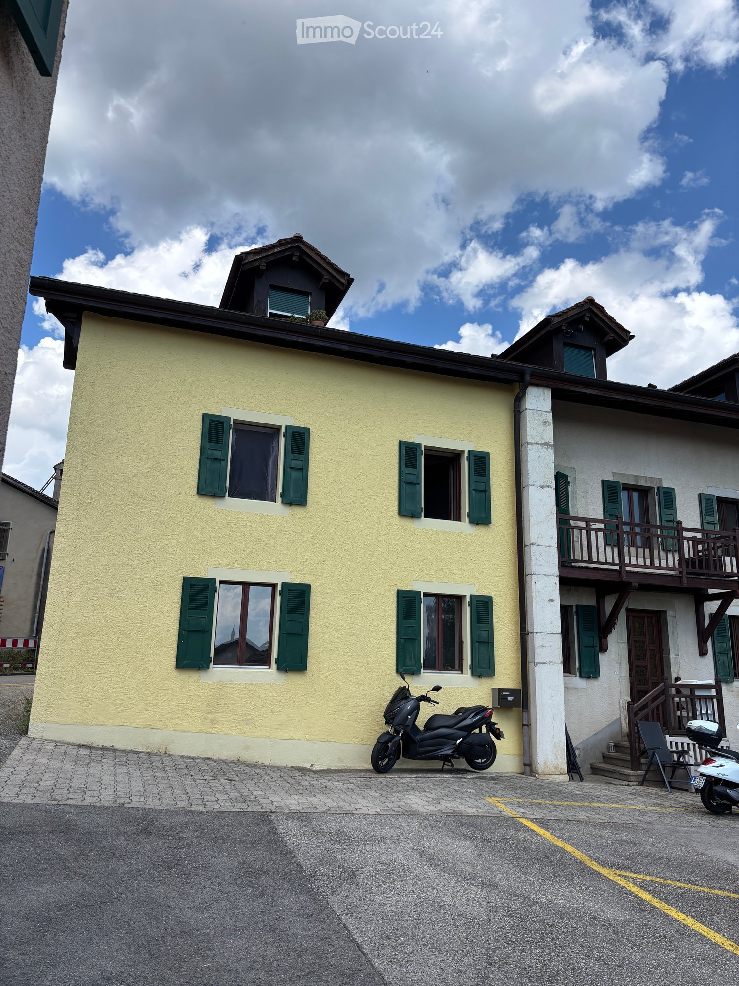Two-story house with yellow walls, green shutters, and a balcony on the upper level. A scooter is parked in front of the house.
