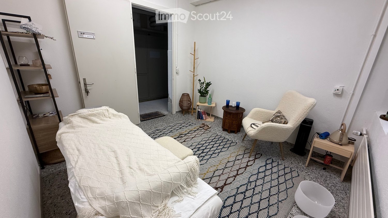 A cozy, well-furnished room with a sofa covered in a white blanket, a white armchair, a small wooden table, a shelf with books and decorative items, a potted plant, and a radiator.