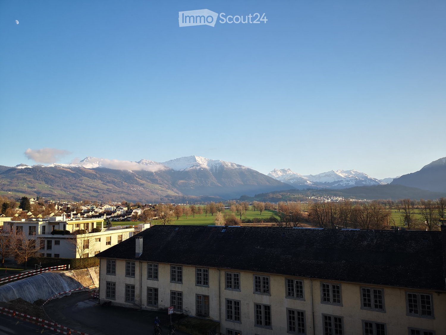 View of a multi-story building with a dark roof and multiple windows, surrounded by a town with visible mountains in the background.