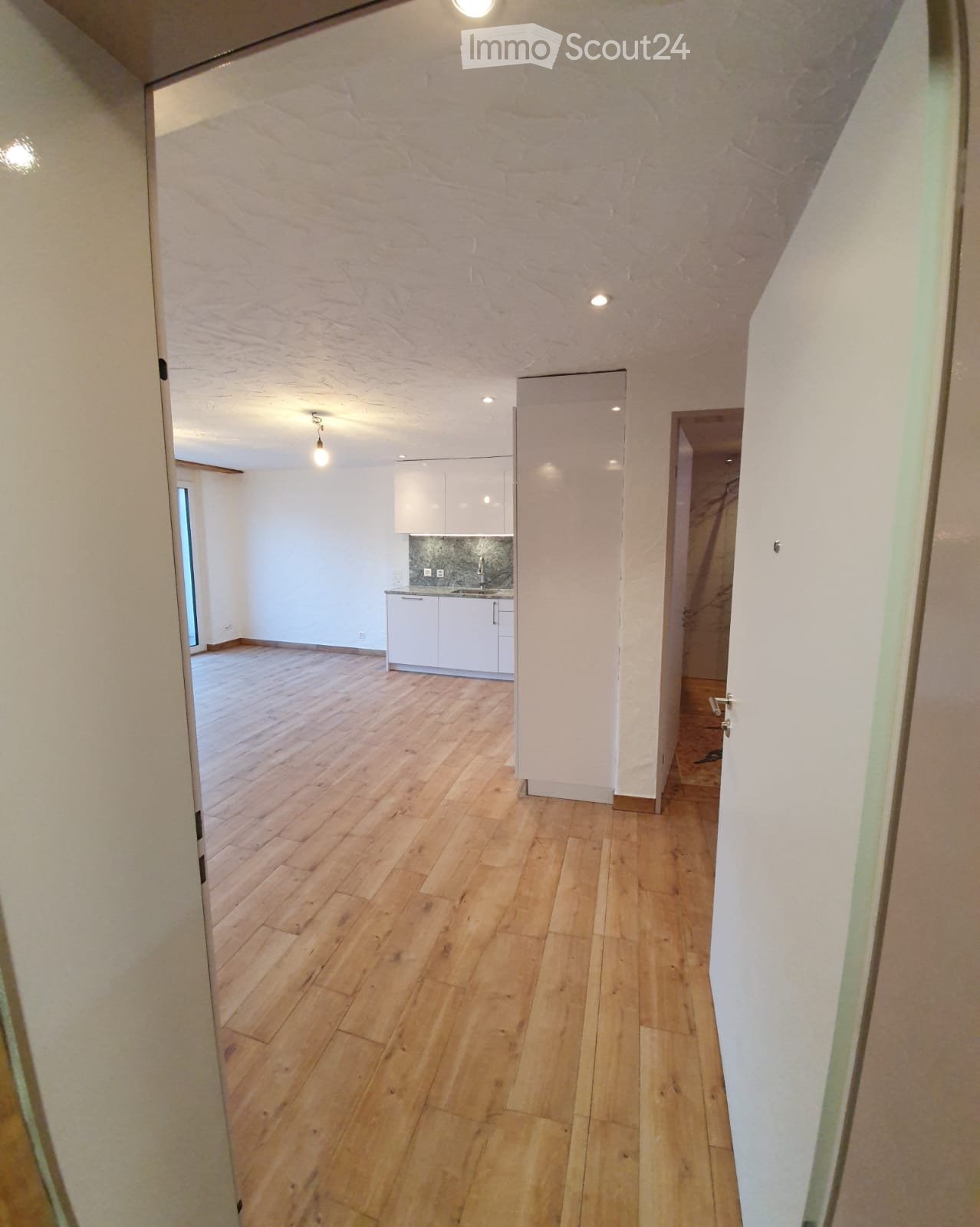 Empty hallway with wooden flooring and white walls, leading to a small kitchen area with white cabinets and a sink.