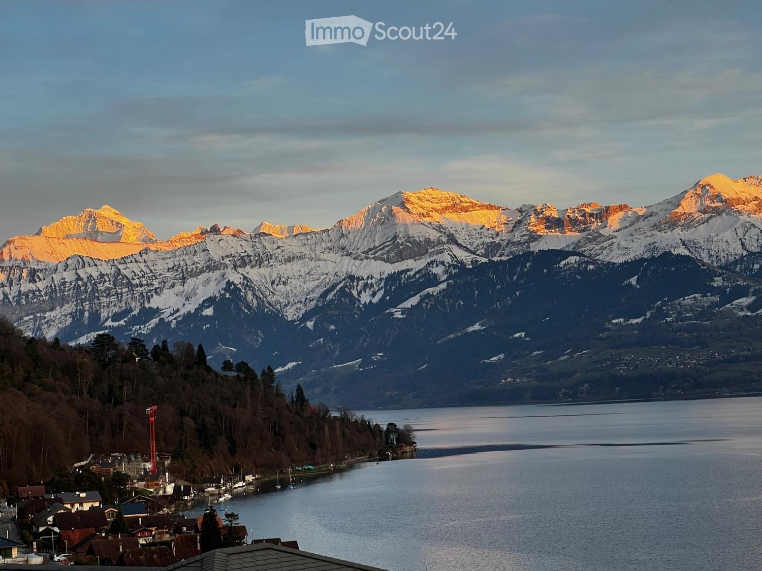 Panoramic view of snow-capped mountains and a calm lake, with a small village on the shore.