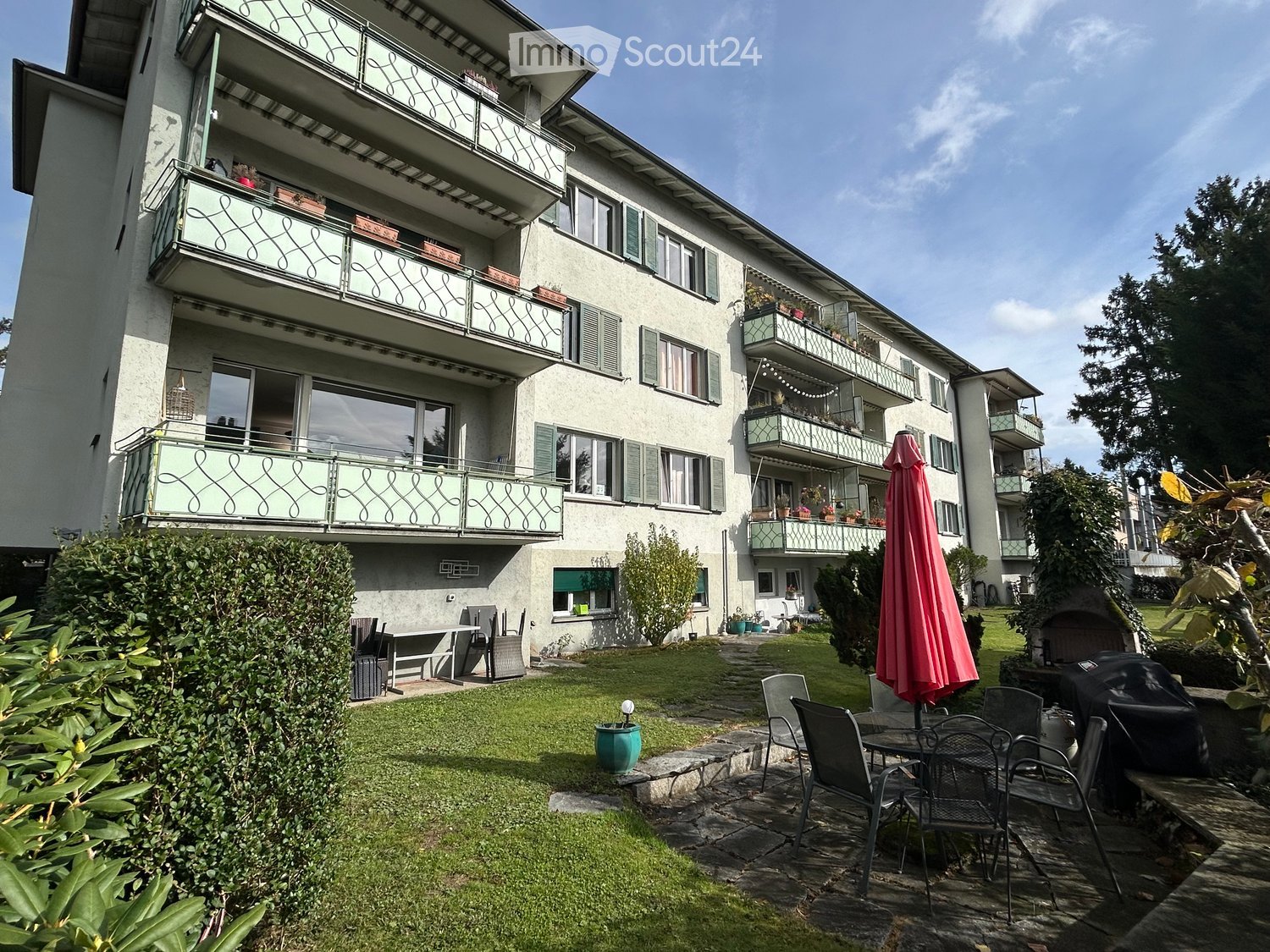 Garden area with a patio, outdoor furniture, and a closed umbrella, adjacent to a multi-story building with balconies.
