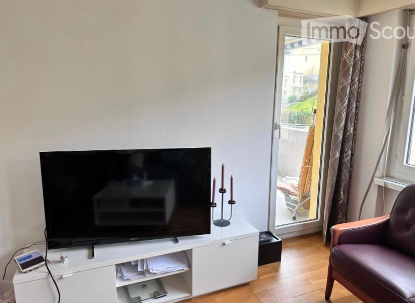 A modern living room with a flat-screen TV on a white cabinet, a purple armchair, and a candle holder on the table. The room has wooden flooring and large windows with curtains.