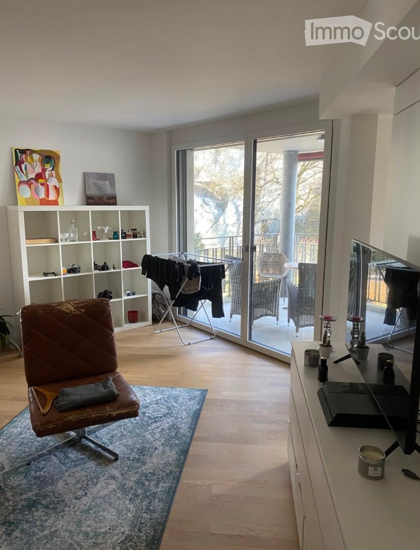 Modern living room with wooden flooring, large sliding glass doors leading to a balcony, a brown leather sofa, a white shelving unit, and a small table with candles.