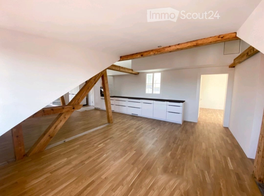An unfurnished attic room with wooden beams, wooden flooring, and white walls. A doorway leads to a kitchen area with white cabinets and a black countertop.