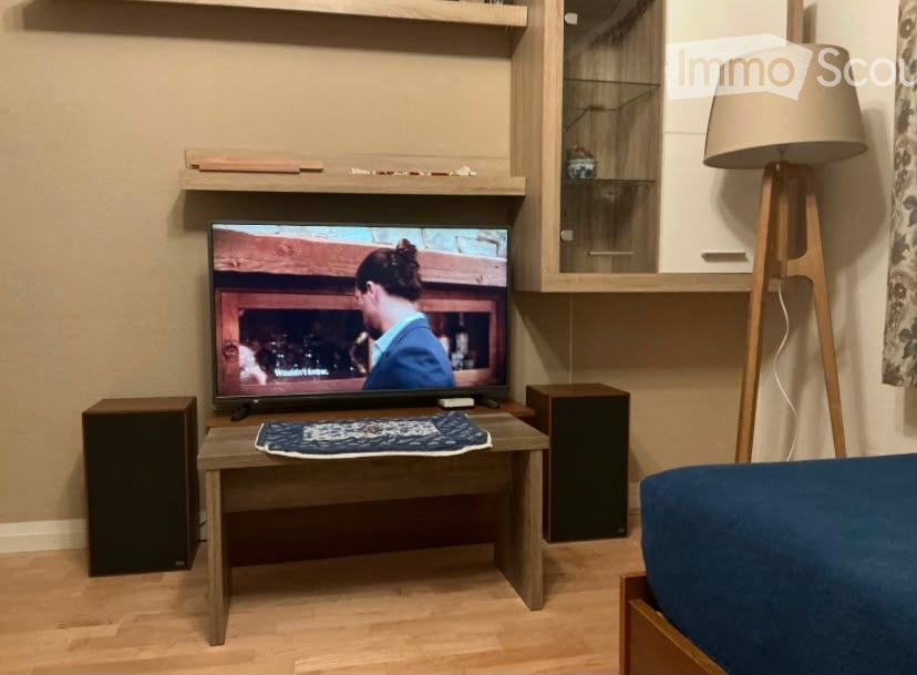 A modern living room with a flat-screen TV on a wooden stand, a small wooden coffee table with a decorative rug, a floor lamp, and two black speakers on either side of the TV.