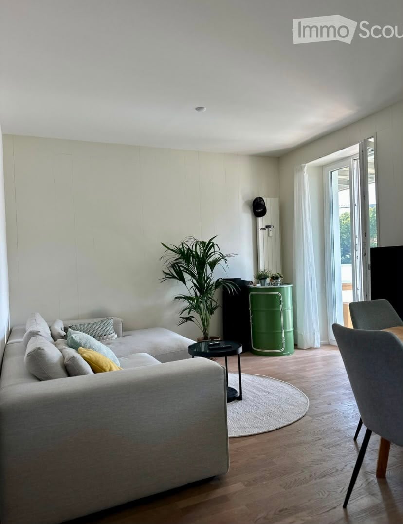 Modern living room with light-colored sofa, coffee table, chairs, a large plant, and a green barrel as a side table. The room features wooden flooring and large windows with white curtains leading to a balcony.