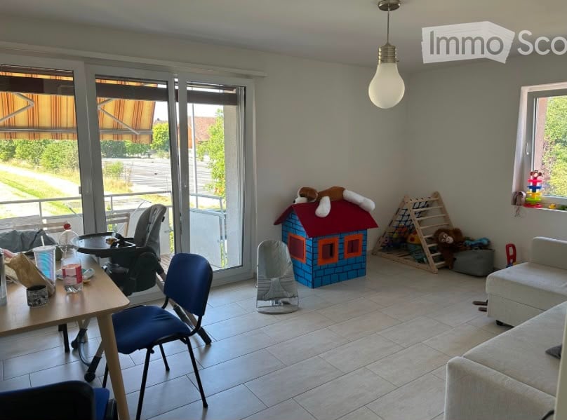 Bright living room with large sliding glass doors leading to a balcony, a sofa, a small table with chairs, a baby chair, a toy house, and various toys including a ladder and stuffed animals.