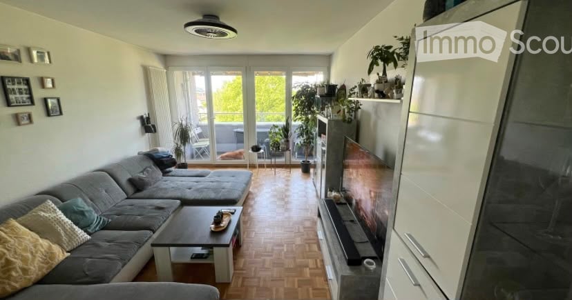 Modern living room with grey sectional sofa, coffee table, TV, and balcony access. Features wooden flooring, white walls, and decorative plants.
