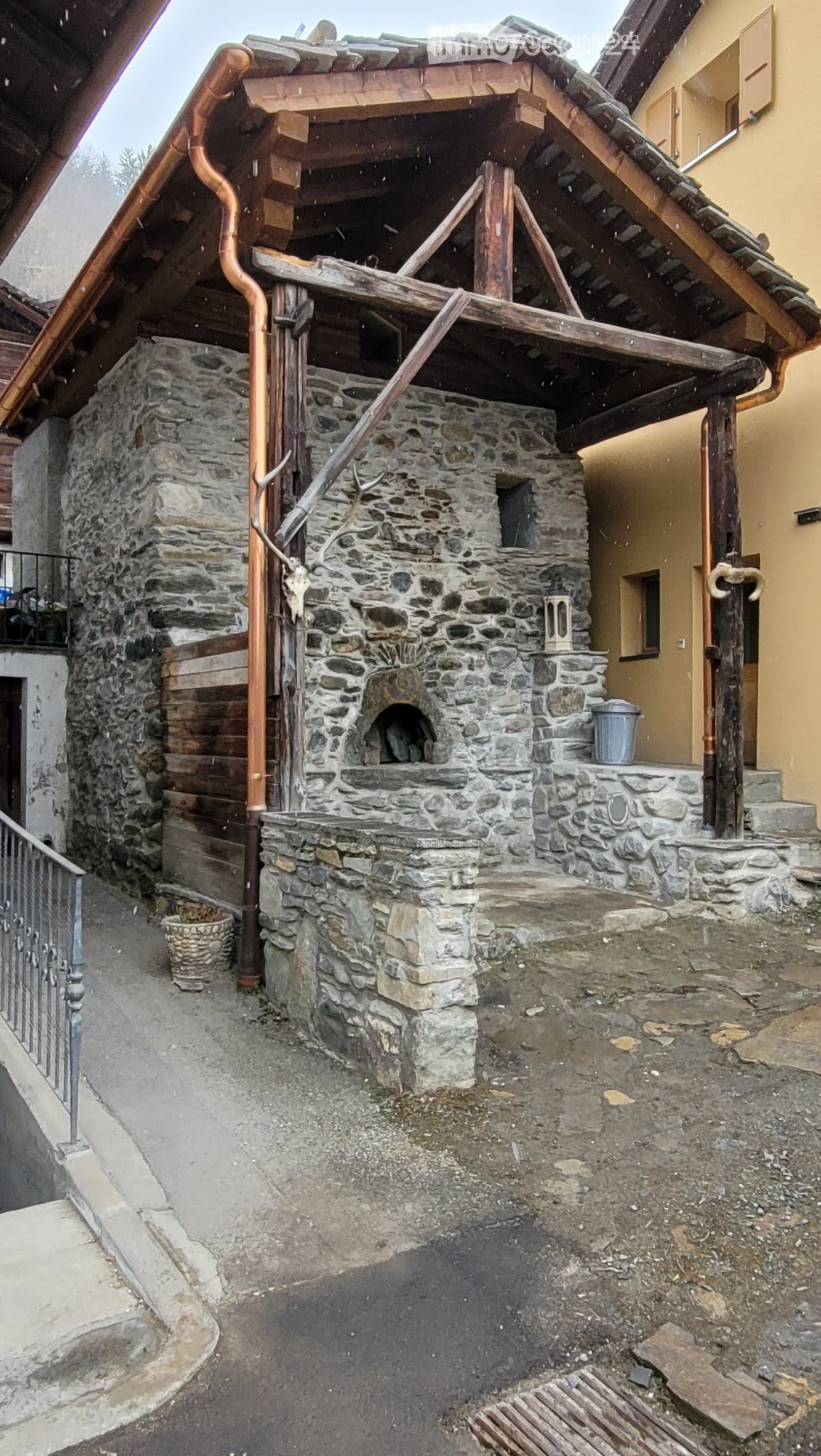 Stone outdoor structure with a raised platform, wooden beams, and a stone oven. The area includes a metal railing, a small basket, and a metal container.