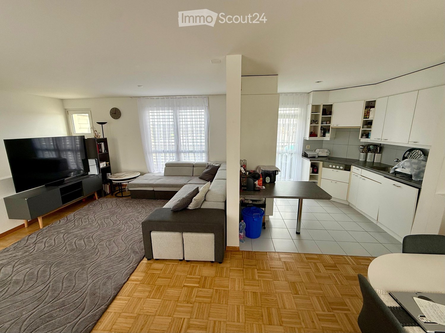 Open-plan living and dining area with a grey sectional sofa, coffee table, TV stand, and a small dining table with chairs. The kitchen features white cabinets, a stove, sink, and fridge.